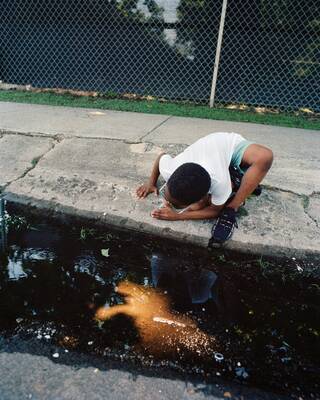 Boy crouched on the curb, looking into a puddle of liquid and seeing a reflection. There is also a bright orange reflection