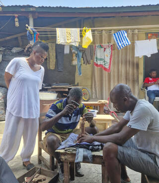 Woman standing next to two men sat down working with hammers on a piece of metal