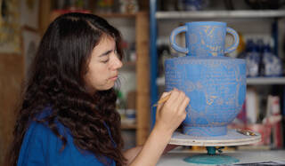 woman with long dark hair sat in front of a large blue and white pot scraping words onto it with a pen-like tool