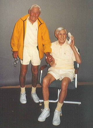 Photograph of an elderly man wearing white t-shirt and shorts and a yellow jacket next to a seated waxwork model of himself 
