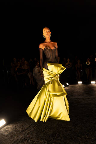 Model on a dark catwalk with floor spotlights wearing black lacy top and yellow skirt with large bow