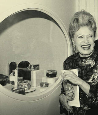 Black and white photograph of a woman smiling in front of a display case of small boxes