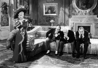 Black and white still image from a movie with three seated men in suits and a woman standing against a chaise longue