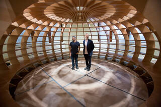 Photograph of two men stood inside a round wooden structure casting shadows on the floor
