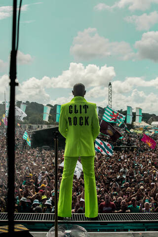 Back of a woman in a green suit stood on a stage in front of a crowd of people outdoors