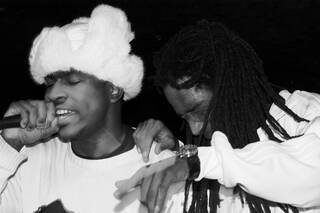 Black and white photo of a man singing with a white fur hat on and another man looking down with dreadlocked hair