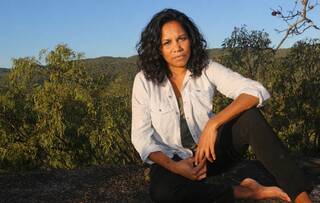 Woman sat outside barefoot on the ground in white jacket and black trousers with backdrop of trees and blue sky