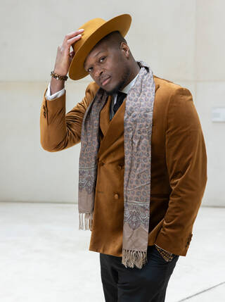 Photo of a man standing in a orange/brown jacket and hat with scarf around his shoulders. He has his right hand on the hat.