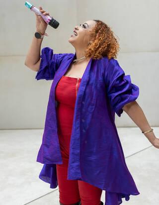 Photo of a woman in a red dress with wavy purple overshirt singing into a microphone