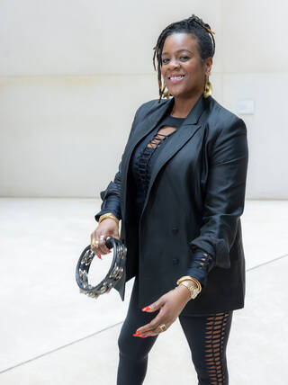 Photo of a standing woman wearing all black with a tambourine in her right hand