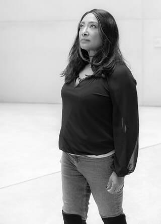 Black and white photo of a standing woman in dark jeans and black top against a pale background