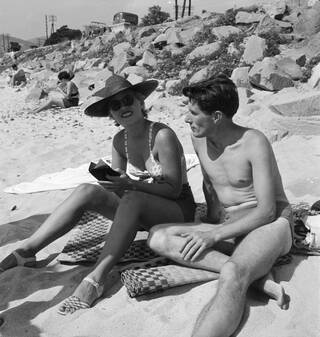 Black and white photograph of a man and woman sat on the beach in swimwear