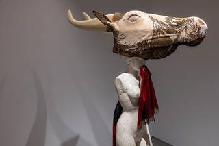 Photograph of a sculpture from the side featuring a human form with large cow skeleton head