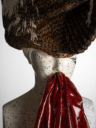 Detail of a sculptural face with section of a brown woven hat and red plastic coming from mouth