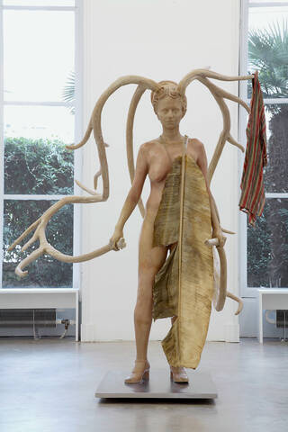 Sculpture of a woman with large antlers with a cloak on one antler and a shield in her hand 