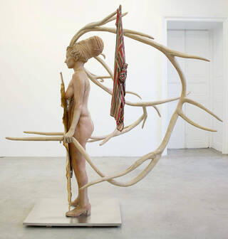 Side view of a sculpture of a woman with large antlers with a cloak hanging from one of them and a shield in her hand