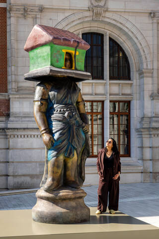Woman looking up at a monumental sculpture of a figure with a house on its head