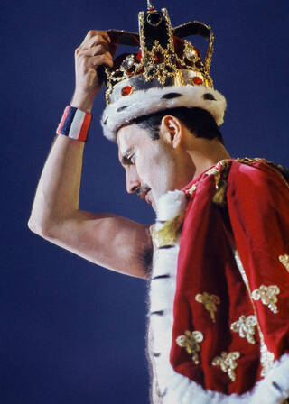 Freddie Mercury on stage wearing a gold crown and red cape against a blue background