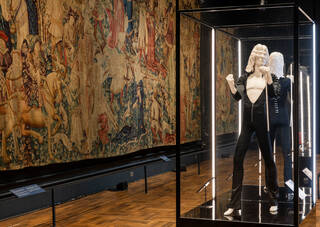 Illuminated glass case with a black costume in it in a room with the walls covered in tapestries