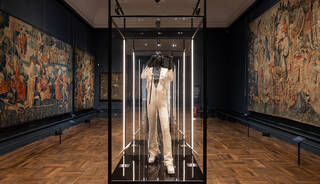 Illuminated glass case with a white costume in it in a room with the walls covered in tapestries