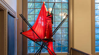 Black and red costume mounted on crossing black scaffolding poles in front of a large window