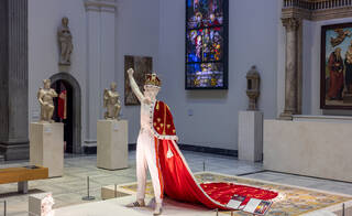 Mannequin wearing gold crown and red cloak with gold embroidery in a room full of sculptures and stained glass panels