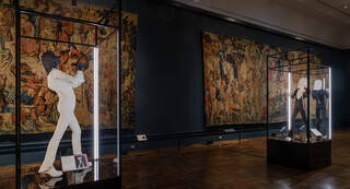 Two illuminated costumes in glass cabinets in a room with tapestries on the walls and a wooden floor